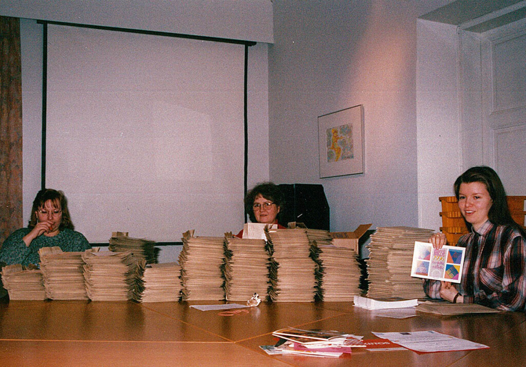 Pöydällä on isot pinot kurssiesitteitä ja kolme naista postittaa niitä.