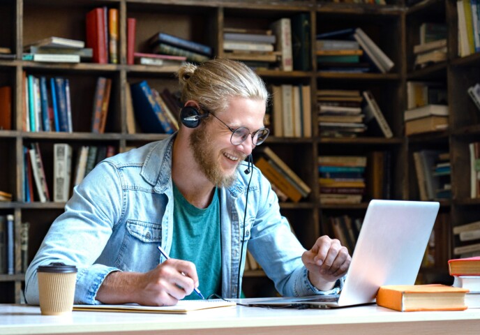 Henkilö istuu tietokoneen ääressä ja hymyilee kuulokkeet päässä.